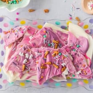 White and pink swirled chocolate bark broken into pieces, topped with colorful sprinkles and small candy shapes, displayed on a polka-dotted tray.