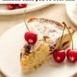 A slice of cherry almond cake topped with powdered sugar and fresh cherries sits on a plate, with additional cherries beside it.