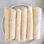 Five rolled air fryer breakfast taquitos are neatly arranged side by side on a white plate, viewed from above on a light gray surface.