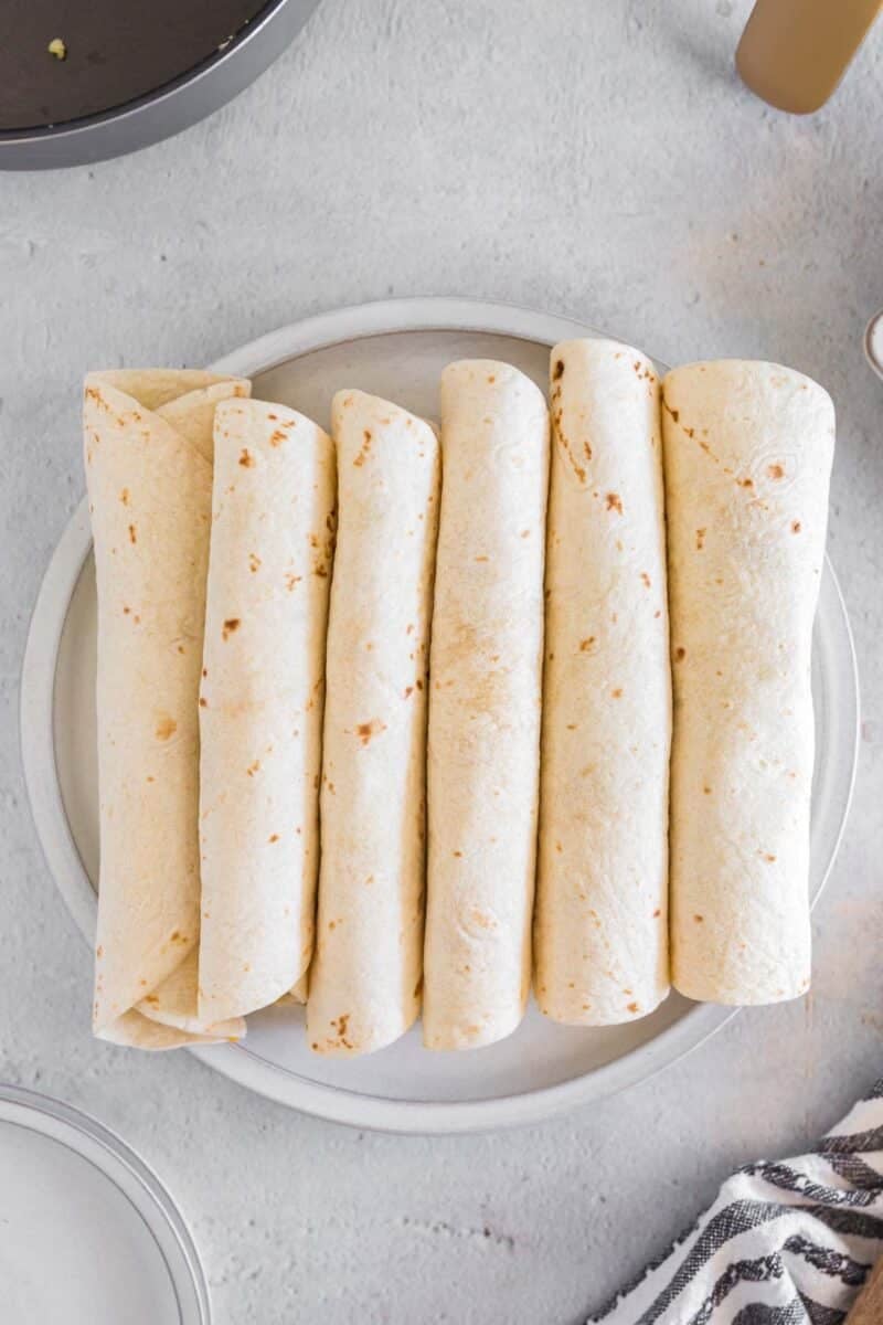 Five rolled air fryer breakfast taquitos are neatly arranged side by side on a white plate, viewed from above on a light gray surface.
