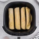 Four air fryer breakfast taquitos are placed in a single layer inside an air fryer basket on a light-colored surface next to a plate and kitchen utensils.