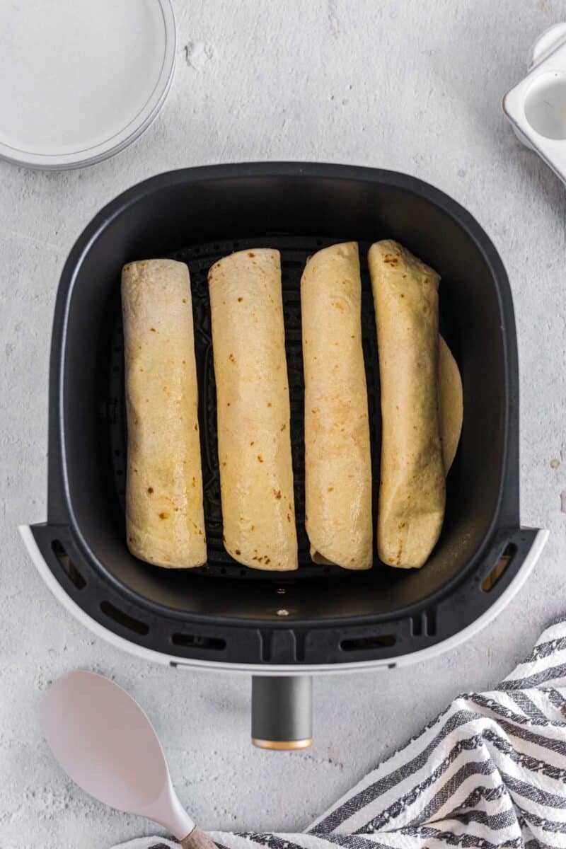 Four air fryer breakfast taquitos are placed in a single layer inside an air fryer basket on a light-colored surface next to a plate and kitchen utensils.