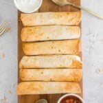 Six baked air fryer breakfast taquitos are arranged in a row on a wooden board, served with bowls of salsa and sour cream, with utensils nearby on a light-colored surface.