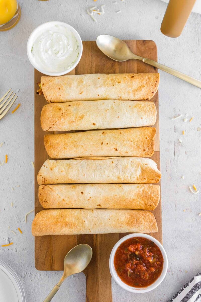 Six baked air fryer breakfast taquitos are arranged in a row on a wooden board, served with bowls of salsa and sour cream, with utensils nearby on a light-colored surface.