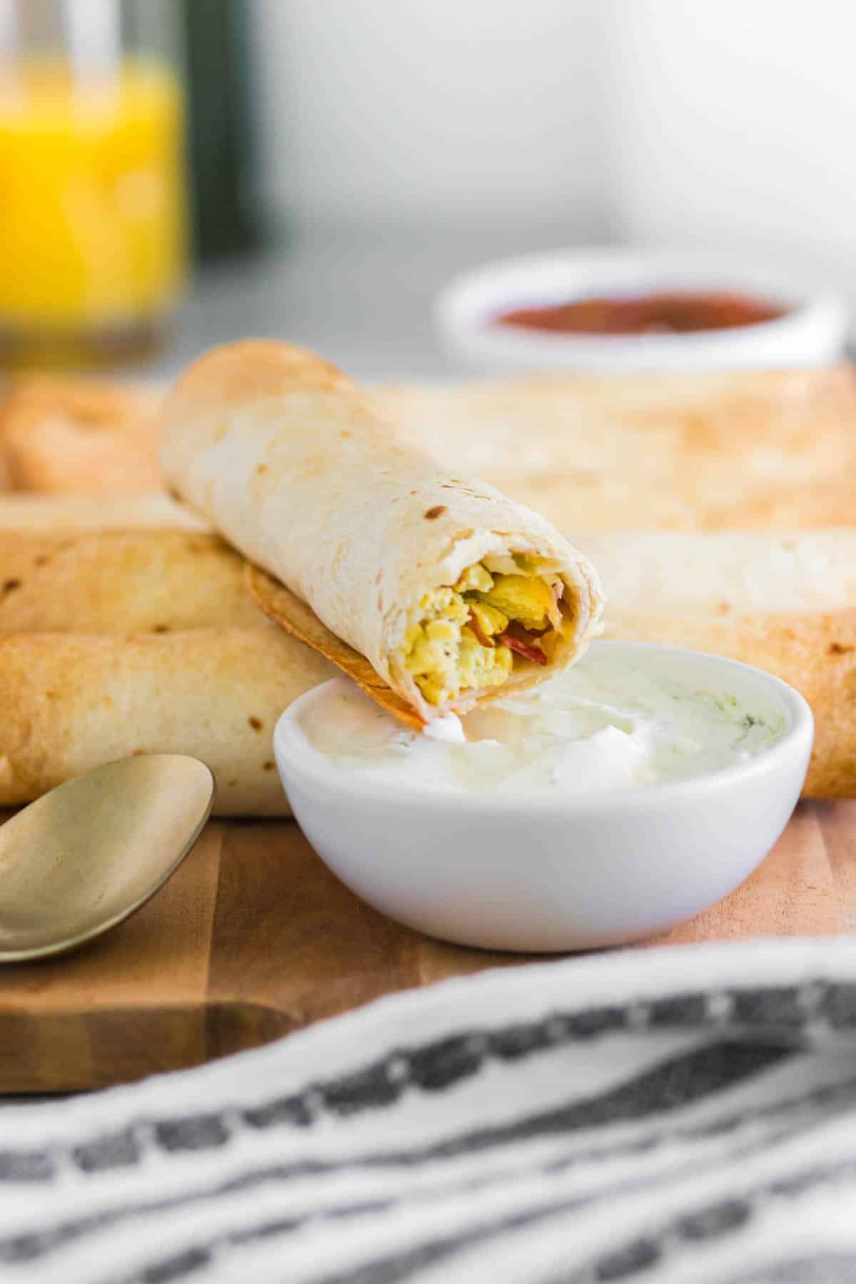 A air fryer breakfast taquito filled with eggs and veggies is dipped in a bowl of sour cream, with more taquitos and a glass of orange juice in the background.