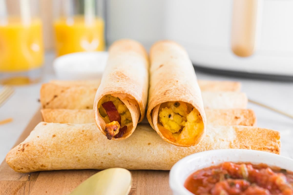 Four air fryer breakfast taquitos filled with scrambled eggs and vegetables are arranged on a wooden board, with salsa and orange juice in the background.