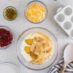 A glass bowl with eggs, milk, and pepper, surrounded by bowls of shredded cheese, chopped bacon, jalapeños, tortillas, a spatula, and a striped towel on a light countertop.