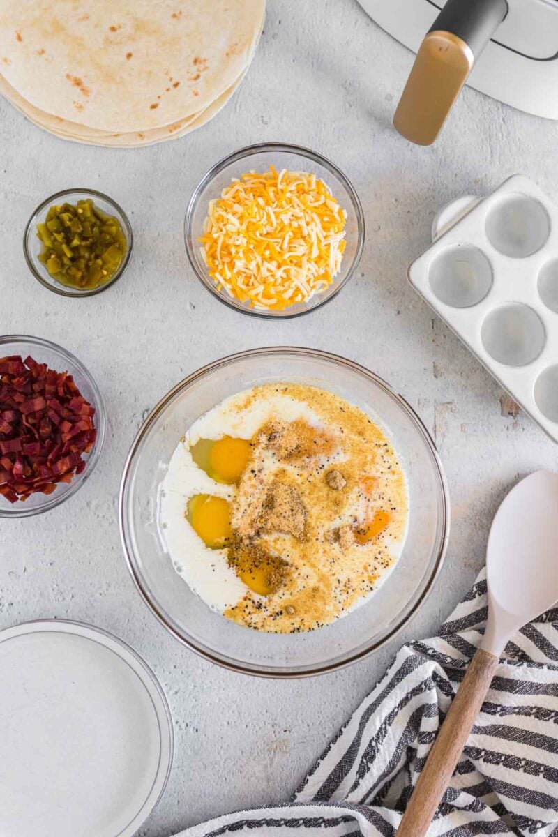A glass bowl with eggs, milk, and pepper, surrounded by bowls of shredded cheese, chopped bacon, jalapeños, tortillas, a spatula, and a striped towel on a light countertop.