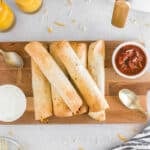 Five air fryer breakfast taquitos on a wooden board, served with bowls of salsa and sour cream, two glasses of orange juice, and gold utensils on a white surface.