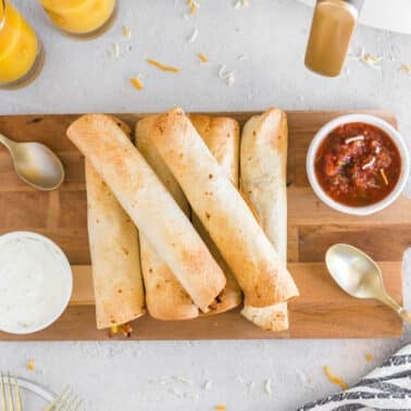 Five air fryer breakfast taquitos on a wooden board, served with bowls of salsa and sour cream, two glasses of orange juice, and gold utensils on a white surface.