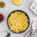 A pan of scrambled eggs on a countertop, surrounded by shredded cheese, chopped jalapeños, bacon bits, tortillas, and kitchen utensils.