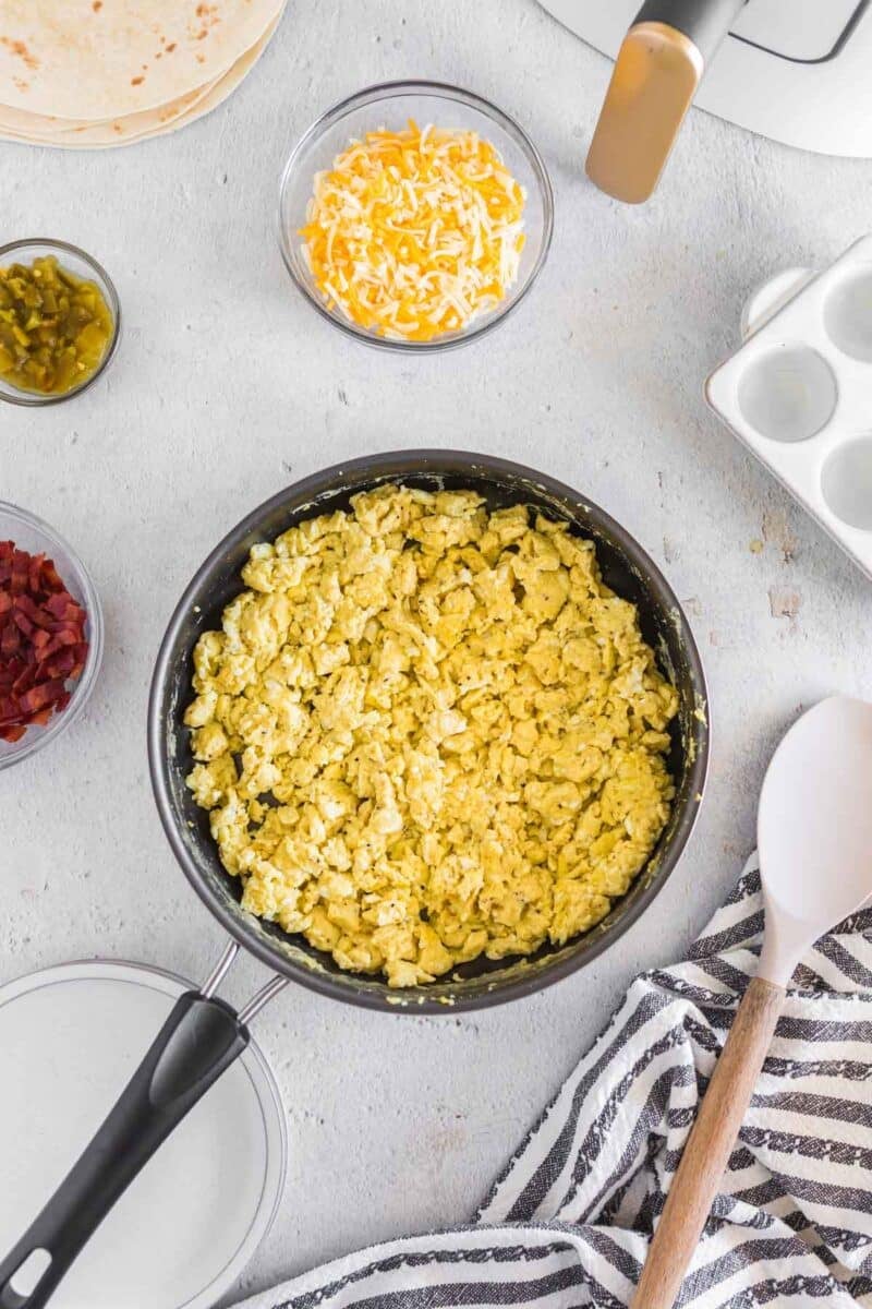 A pan of scrambled eggs on a countertop, surrounded by shredded cheese, chopped jalapeños, bacon bits, tortillas, and kitchen utensils.