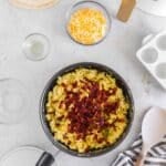 A pan of scrambled eggs topped with chopped bacon sits on a gray surface, surrounded by shredded cheese, tortillas, a striped towel, and an air fryer.