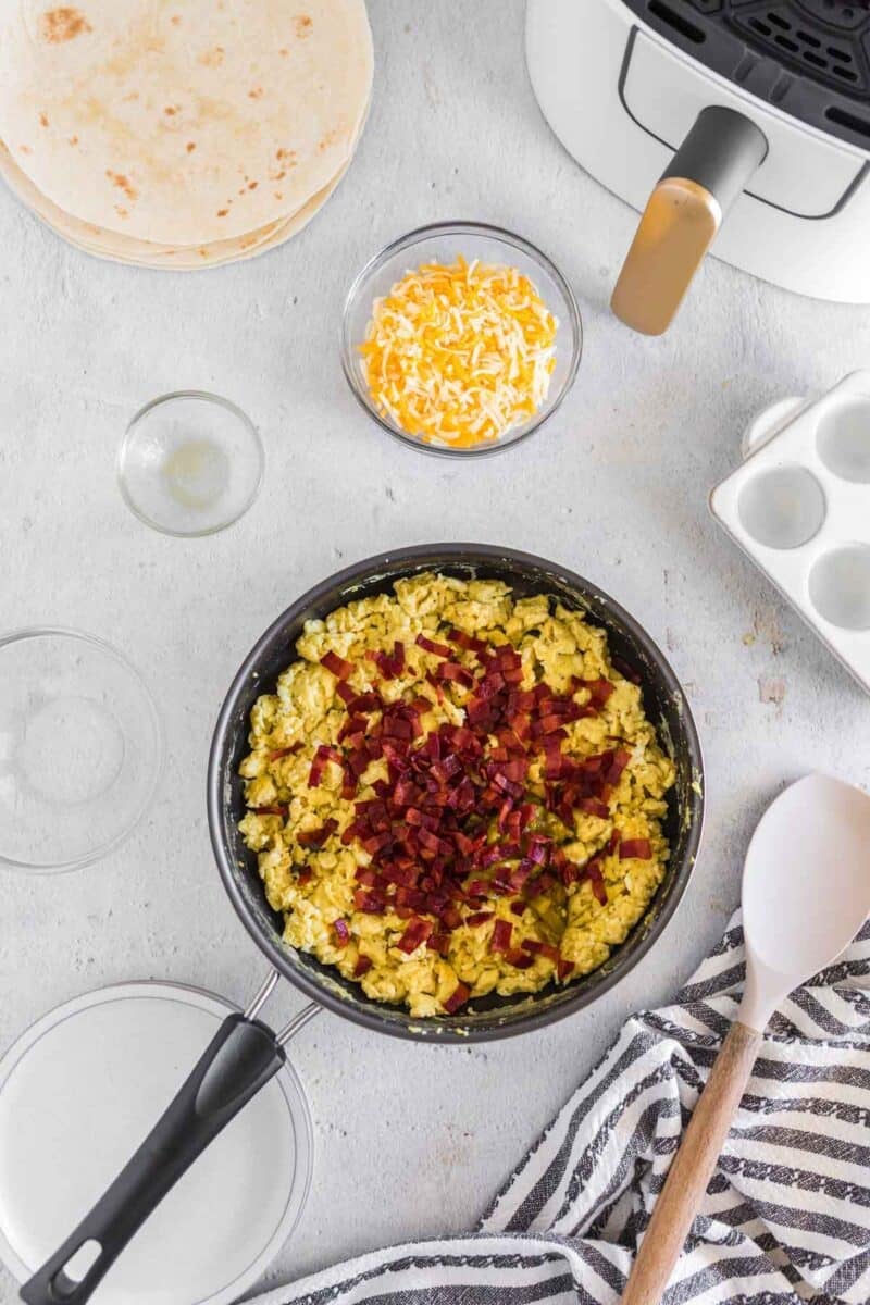 A pan of scrambled eggs topped with chopped bacon sits on a gray surface, surrounded by shredded cheese, tortillas, a striped towel, and an air fryer.
