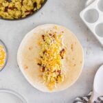 A flour tortilla on a counter is topped with scrambled eggs, bacon bits, and shredded cheese, ready to be wrapped. A skillet, bowl of cheese, and striped towel are nearby.