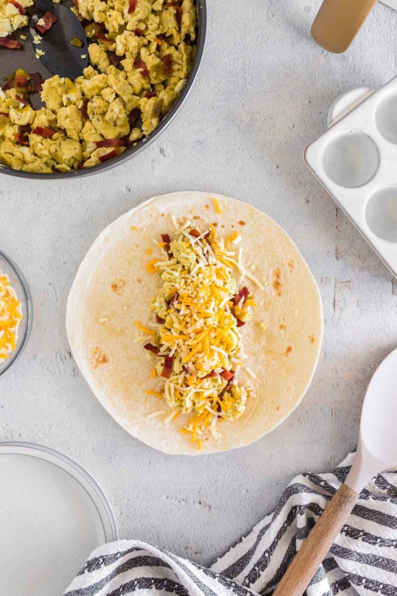 A flour tortilla on a counter is topped with scrambled eggs, bacon bits, and shredded cheese, ready to be wrapped. A skillet, bowl of cheese, and striped towel are nearby.