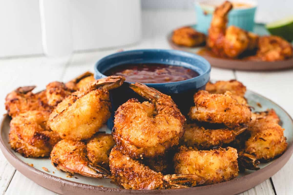 A plate of golden-brown air fryer coconut shrimp is served with a bowl of red dipping sauce; more shrimp are visible in the background.
