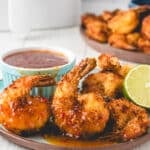 Plate of air fryer coconut shrimp with sauce, a lime half, and extra shrimp in the background, served on a blue plate on a white table.