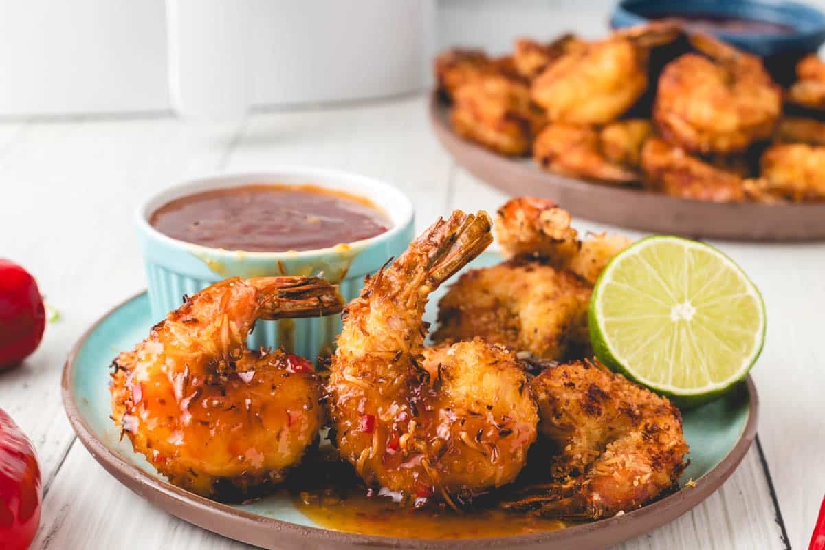 Plate of air fryer coconut shrimp with sauce, a lime half, and extra shrimp in the background, served on a blue plate on a white table.