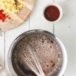 A saucepan with a whisk mixing a brown liquid, next to a cutting board with chopped ginger, garlic, and chili, and small bowls of sauce and cornstarch.