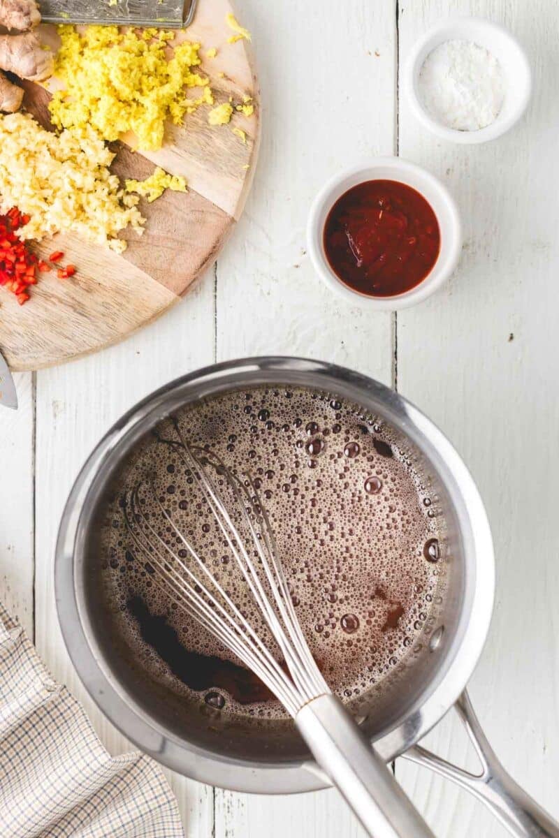 A saucepan with a whisk mixing a brown liquid, next to a cutting board with chopped ginger, garlic, and chili, and small bowls of sauce and cornstarch.