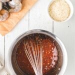 A metal whisk in a saucepan with red sauce, next to a bowl of shredded cheese and raw shrimp on brown paper, on a white wooden surface.