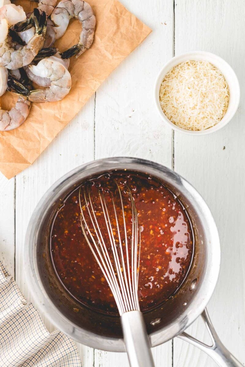 A metal whisk in a saucepan with red sauce, next to a bowl of shredded cheese and raw shrimp on brown paper, on a white wooden surface.