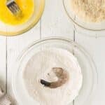 A raw shrimp sits in a bowl of flour, with a bowl of beaten egg and a bowl of shredded coconut nearby on a white wooden surface.