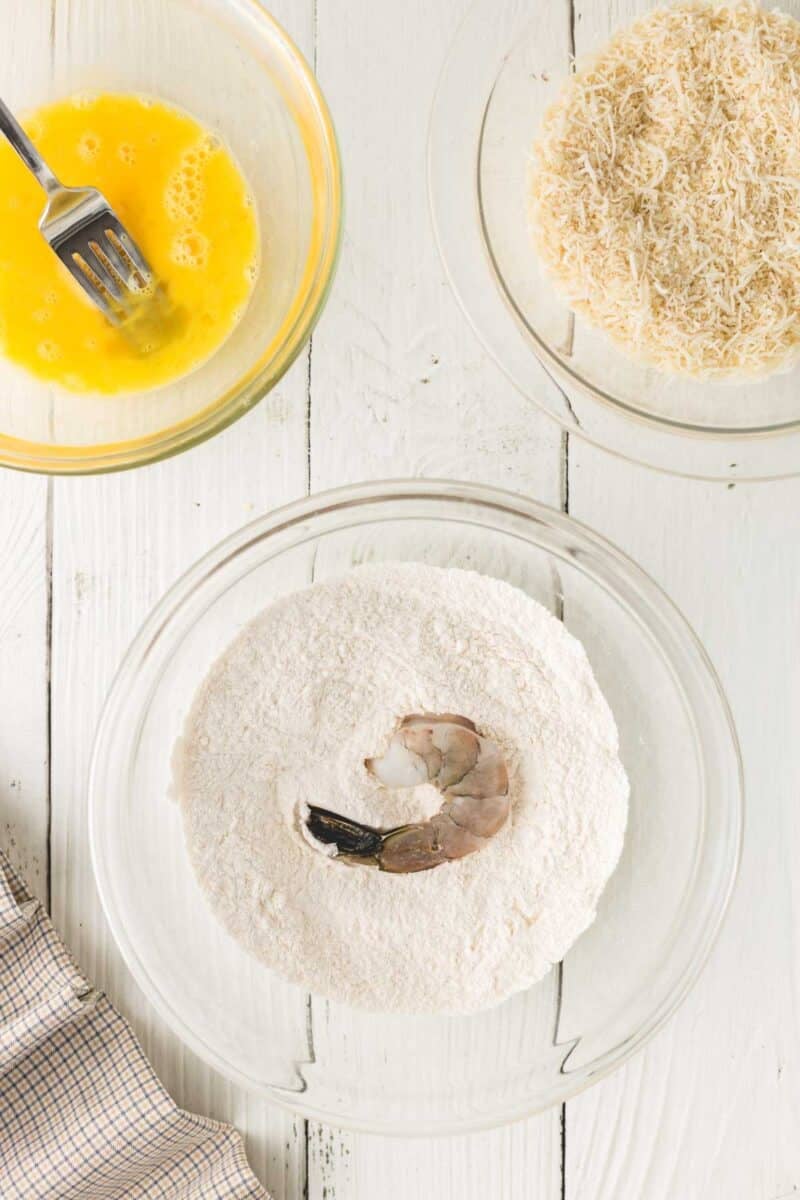 A raw shrimp sits in a bowl of flour, with a bowl of beaten egg and a bowl of shredded coconut nearby on a white wooden surface.