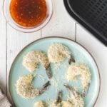 A plate of uncooked, breaded shrimp arranged in a circle, next to an air fryer basket and a bowl of orange dipping sauce on a white wooden table.