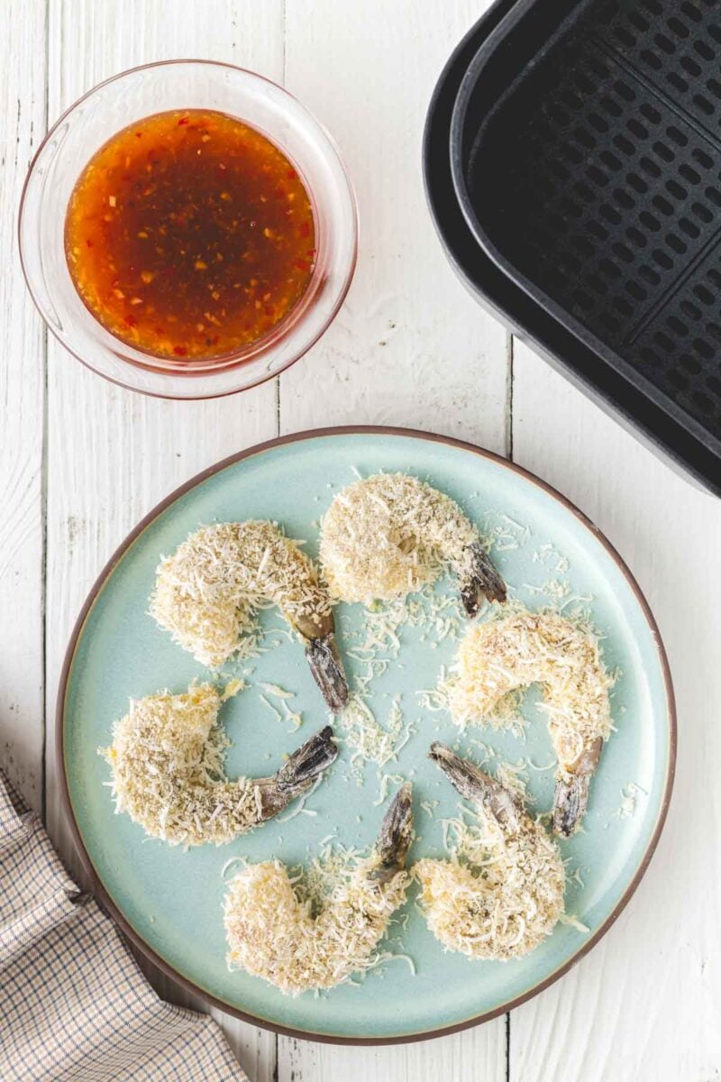 A plate of uncooked, breaded shrimp arranged in a circle, next to an air fryer basket and a bowl of orange dipping sauce on a white wooden table.