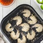 Breaded shrimp in an air fryer basket, with a bowl of dipping sauce, two limes, and a bottle of avocado oil arranged on a white wooden surface.