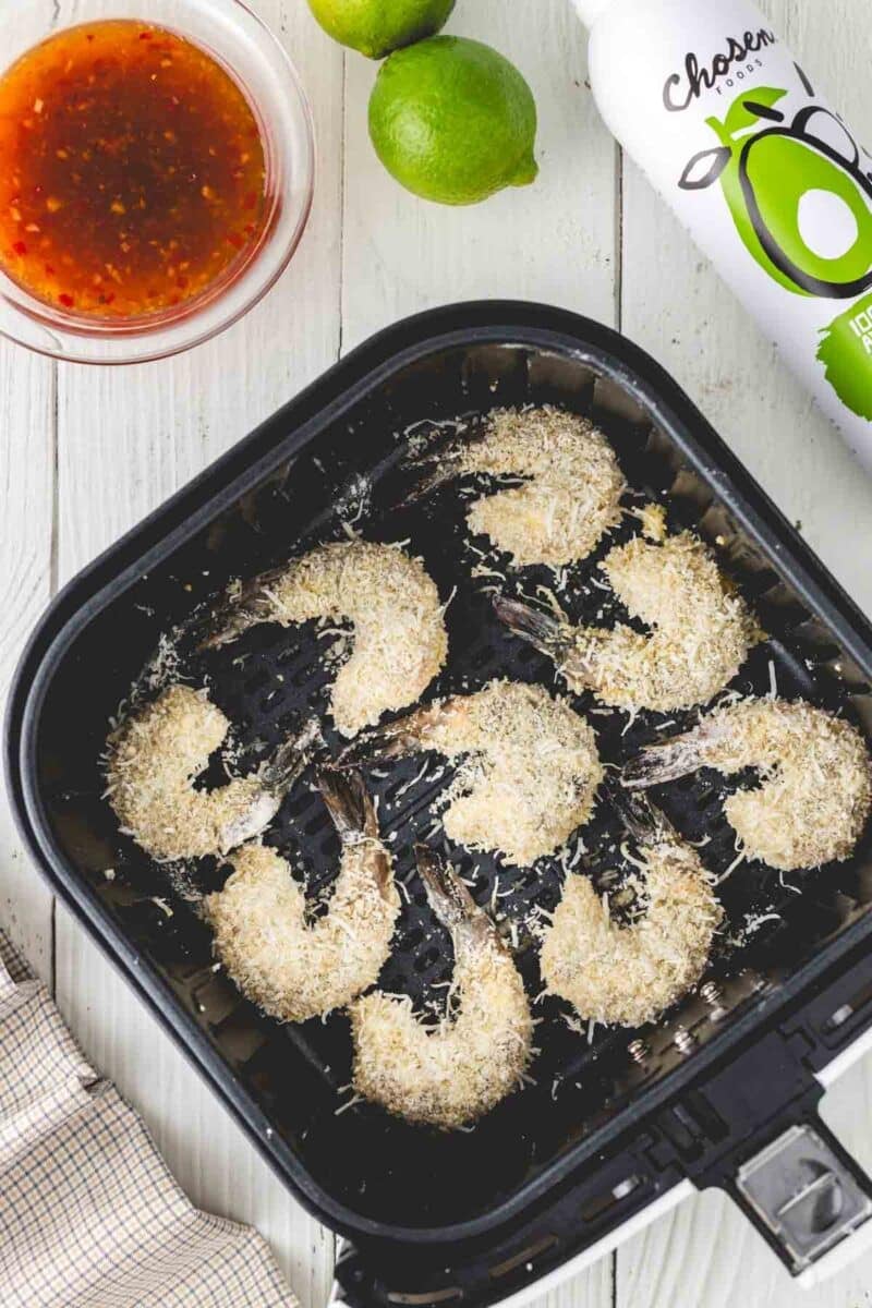 Breaded shrimp in an air fryer basket, with a bowl of dipping sauce, two limes, and a bottle of avocado oil arranged on a white wooden surface.