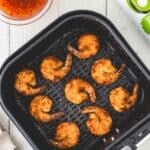 Eight breaded shrimp in an air fryer basket, with a bowl of dipping sauce, two limes, and a bottle of oil on a white surface.