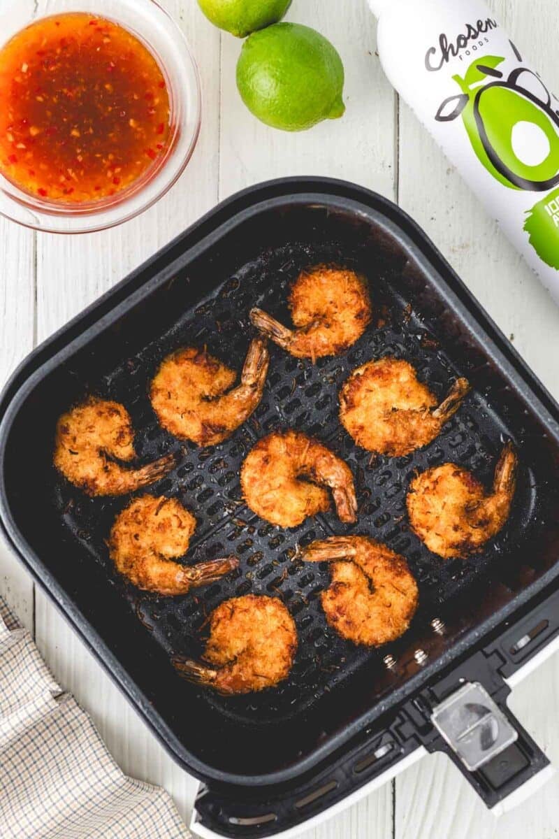 Eight breaded shrimp in an air fryer basket, with a bowl of dipping sauce, two limes, and a bottle of oil on a white surface.