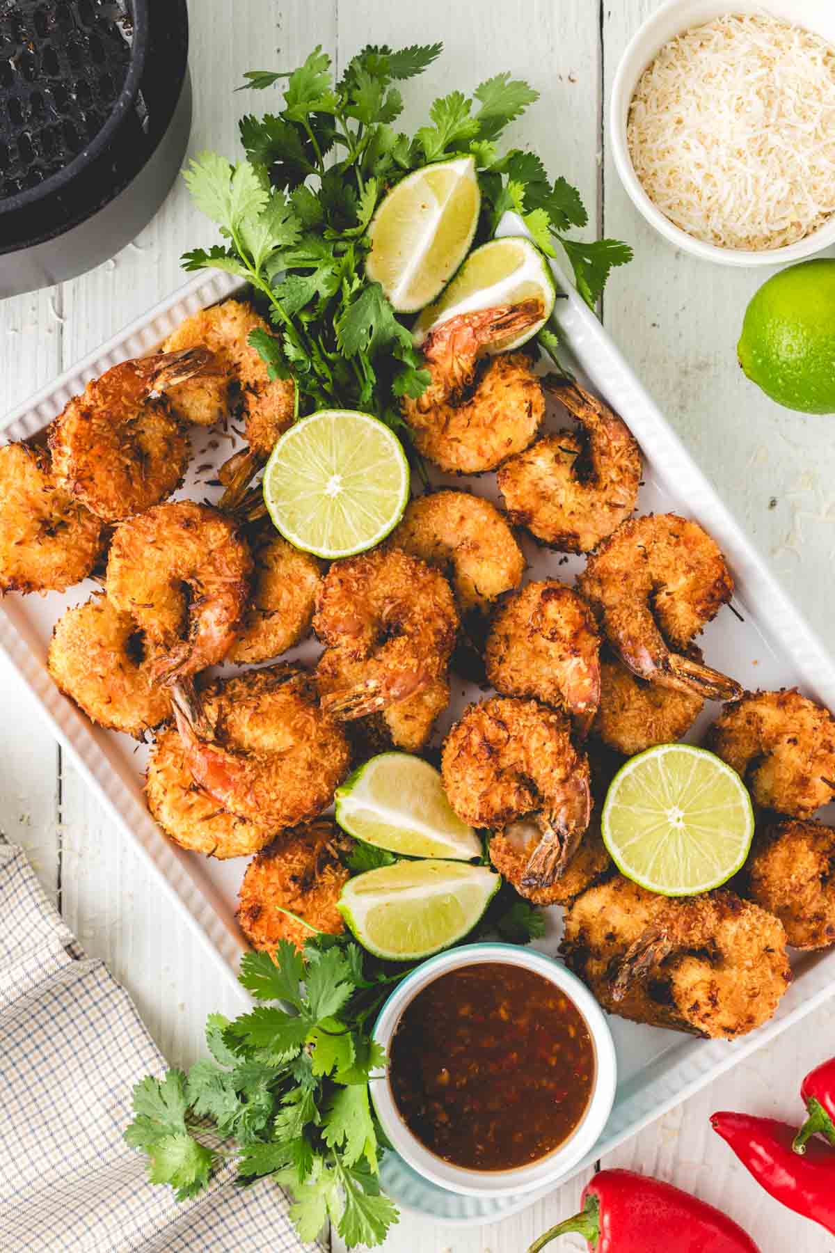 A plate of breaded fried shrimp garnished with lime wedges and cilantro, served with dipping sauce; rice, a lime, and red chili peppers are nearby.