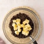 A clear glass bowl with chopped dark chocolate and cubed butter sits on a woven mat, with a saucepan handle visible at the edge.