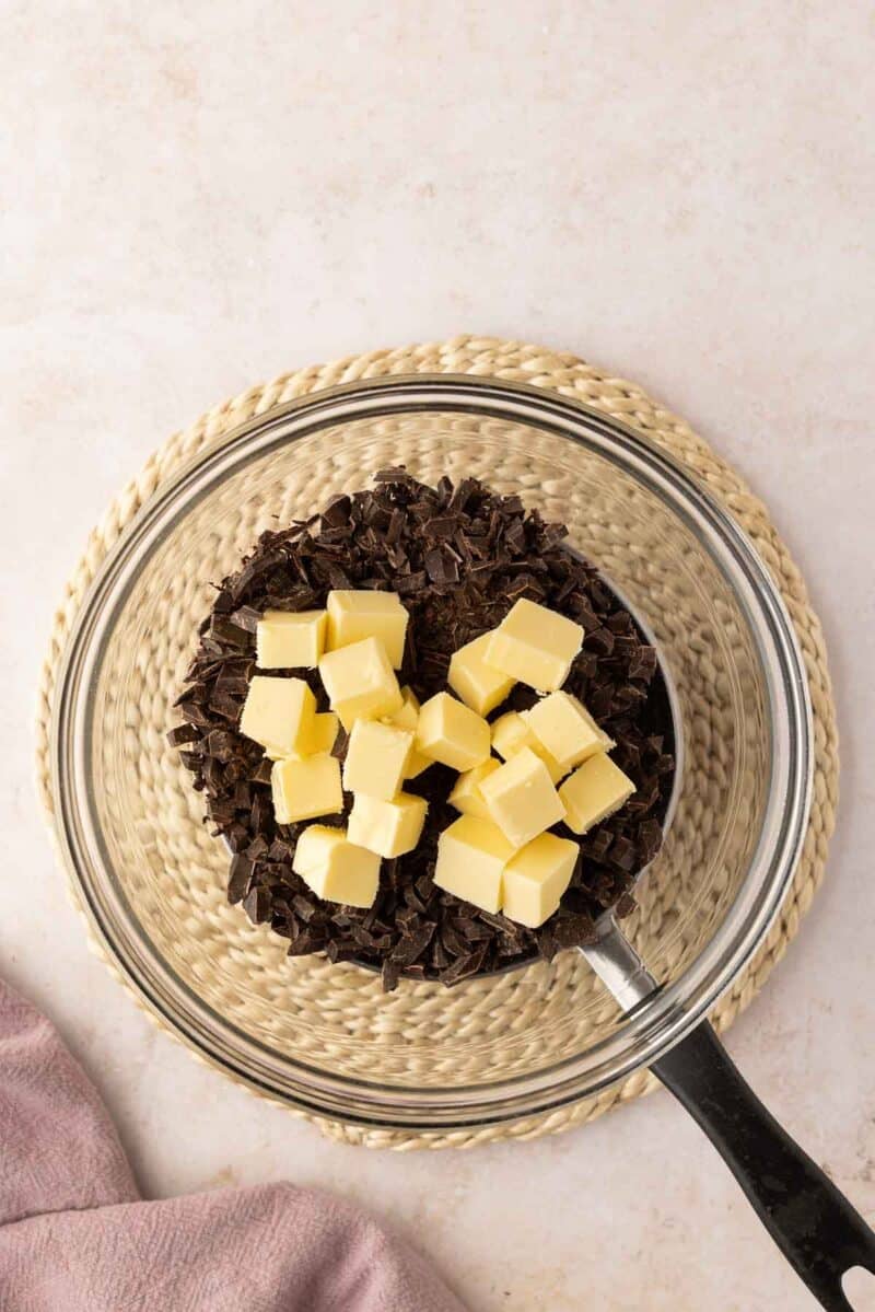 A clear glass bowl with chopped dark chocolate and cubed butter sits on a woven mat, with a saucepan handle visible at the edge.