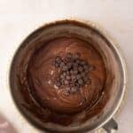 A metal mixing bowl with chocolate batter and chocolate chips on top, placed on a light surface with a mauve cloth partially visible in the corner.