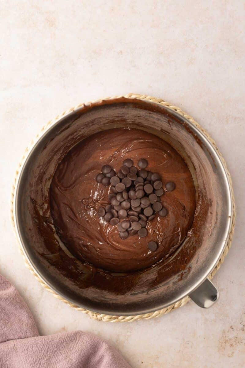 A metal mixing bowl with chocolate batter and chocolate chips on top, placed on a light surface with a mauve cloth partially visible in the corner.
