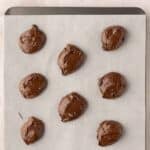 Nine scoops of chocolate cookie dough are placed on a parchment-lined baking sheet, ready to be baked.