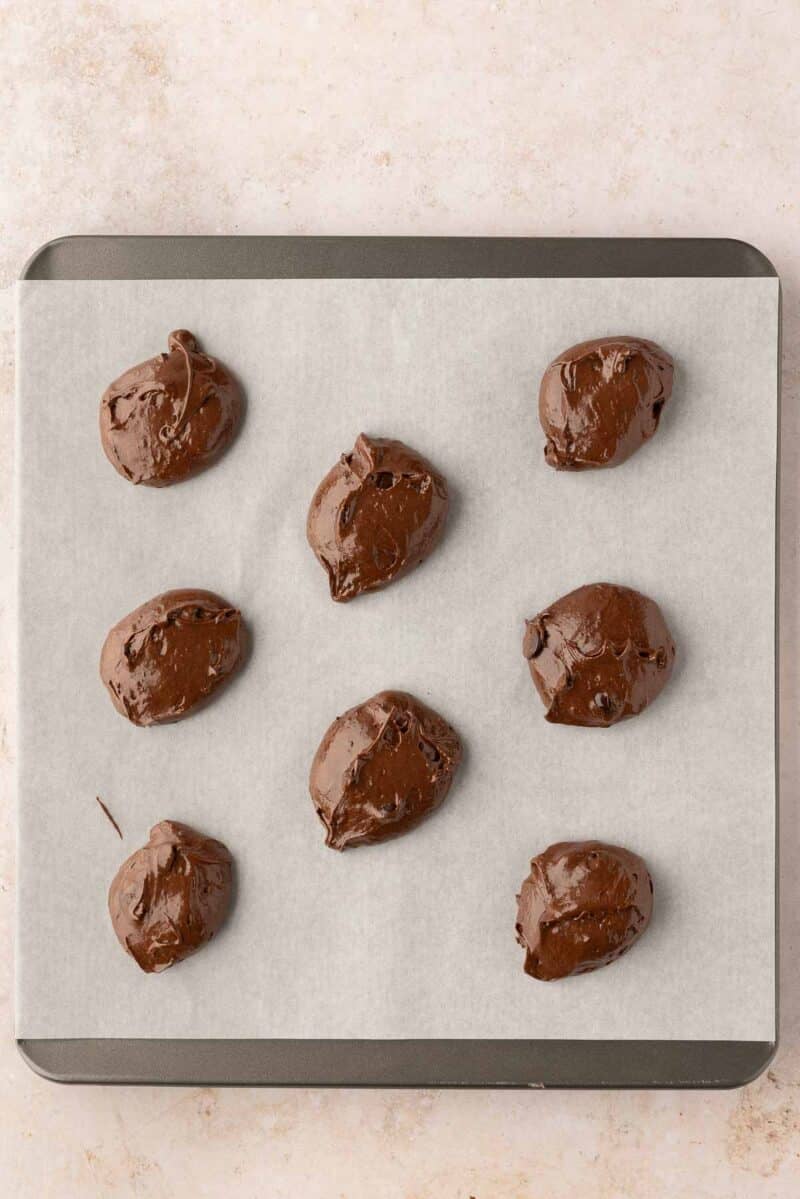 Nine scoops of chocolate cookie dough are placed on a parchment-lined baking sheet, ready to be baked.