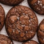 Close-up of several brownie cookies with a cracked surface, sprinkled with flaky sea salt, arranged on a sheet of white parchment paper.