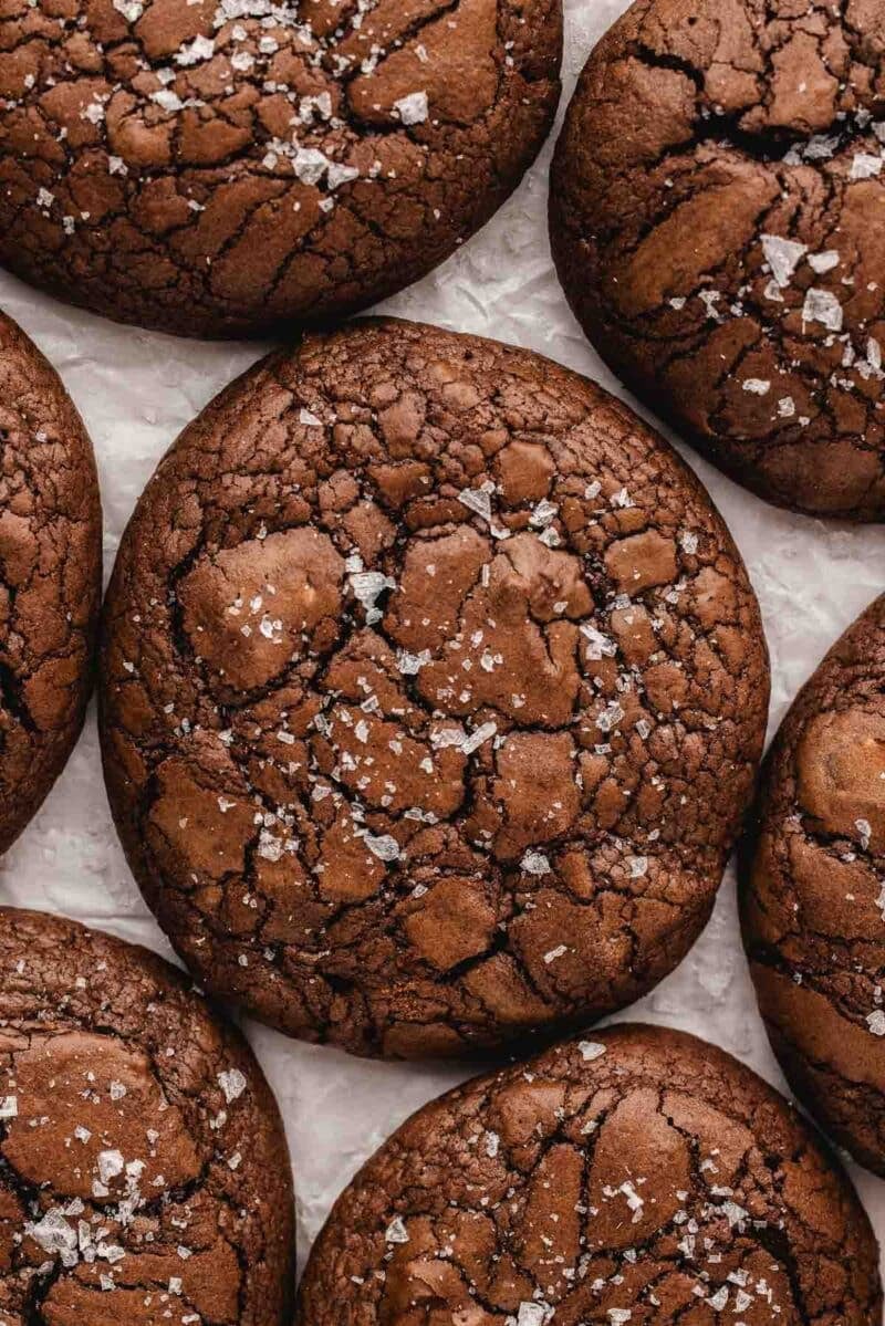 Close-up of several brownie cookies with a cracked surface, sprinkled with flaky sea salt, arranged on a sheet of white parchment paper.