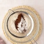 A metal sifter with flour, cocoa powder, and baking powder sits on a glass bowl, placed on a woven mat with a pink cloth nearby.