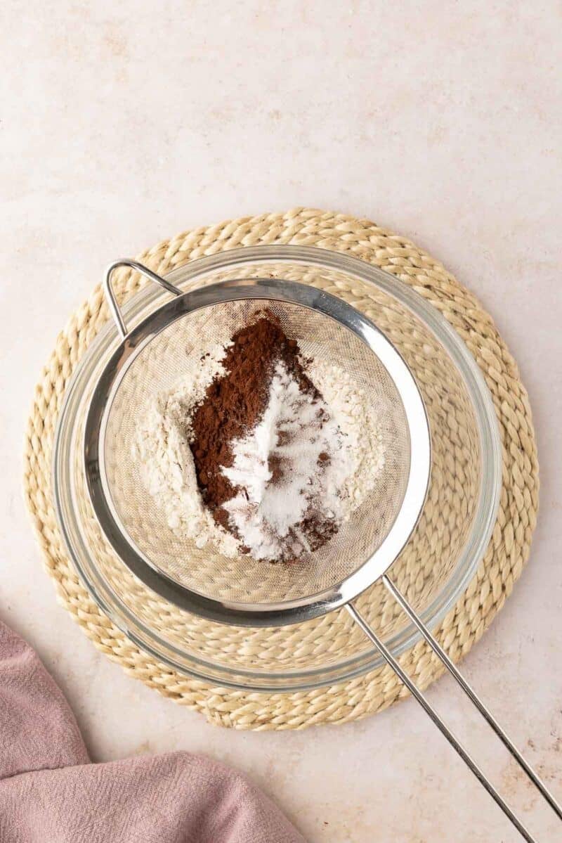 A metal sifter with flour, cocoa powder, and baking powder sits on a glass bowl, placed on a woven mat with a pink cloth nearby.
