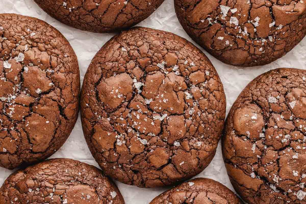 Close-up of several cracked brownie cookies sprinkled with coarse sea salt, arranged on parchment paper.