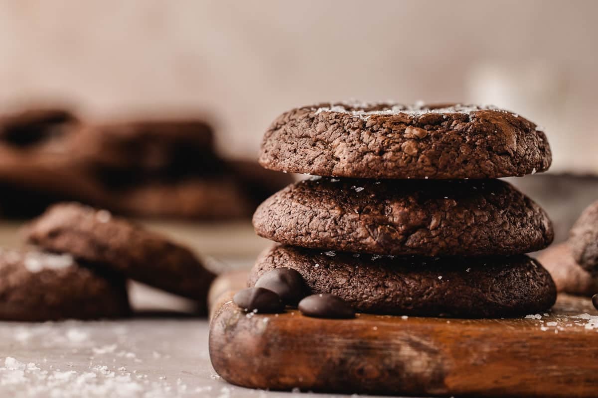 Three brownie cookies stacked on a wooden board with chocolate chips and scattered coarse salt, with more cookies blurred in the background.