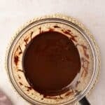 A glass bowl filled with melted chocolate sits on a woven mat, with a saucepan handle visible and a mauve cloth nearby.