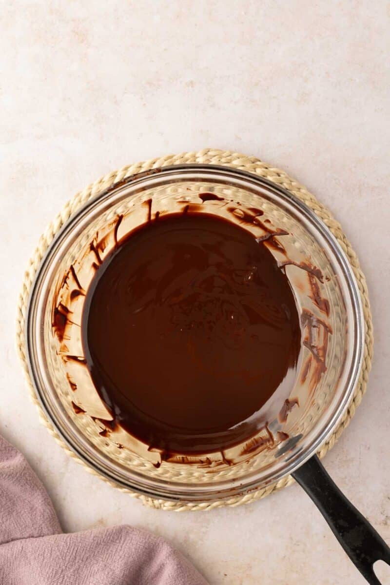 A glass bowl filled with melted chocolate sits on a woven mat, with a saucepan handle visible and a mauve cloth nearby.